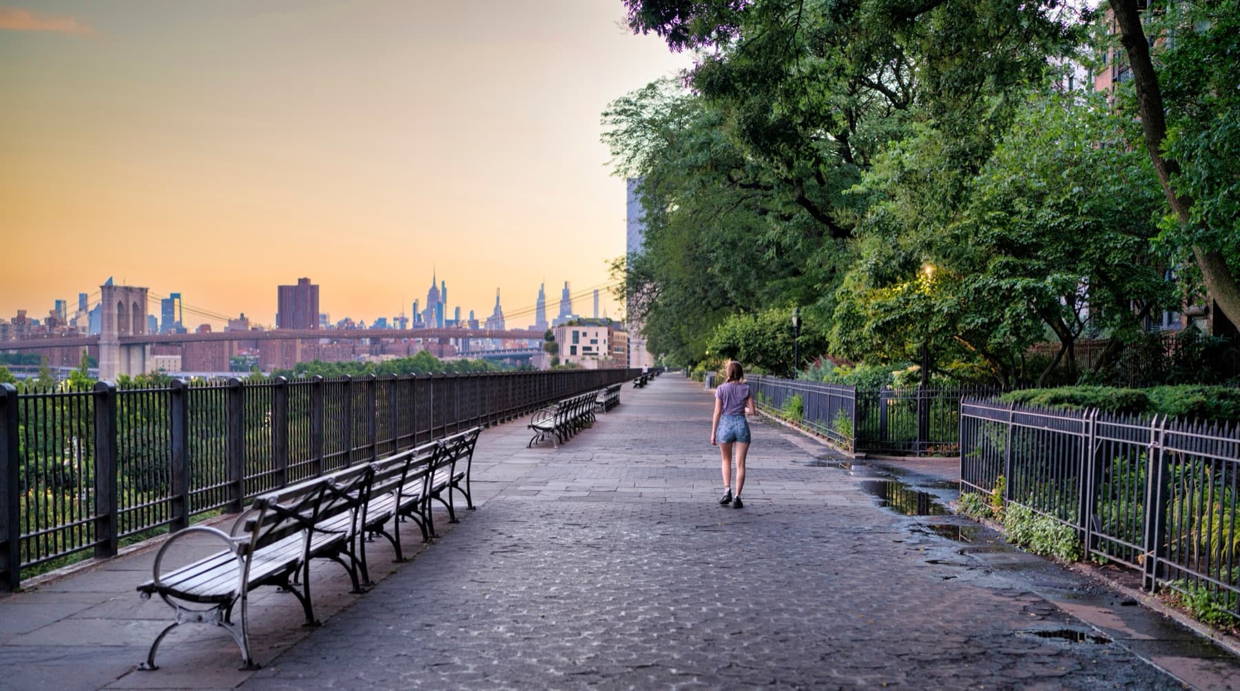 A woman walking along the Brooklyn Heights Promenade at sunset, Manhattan skyline behind her