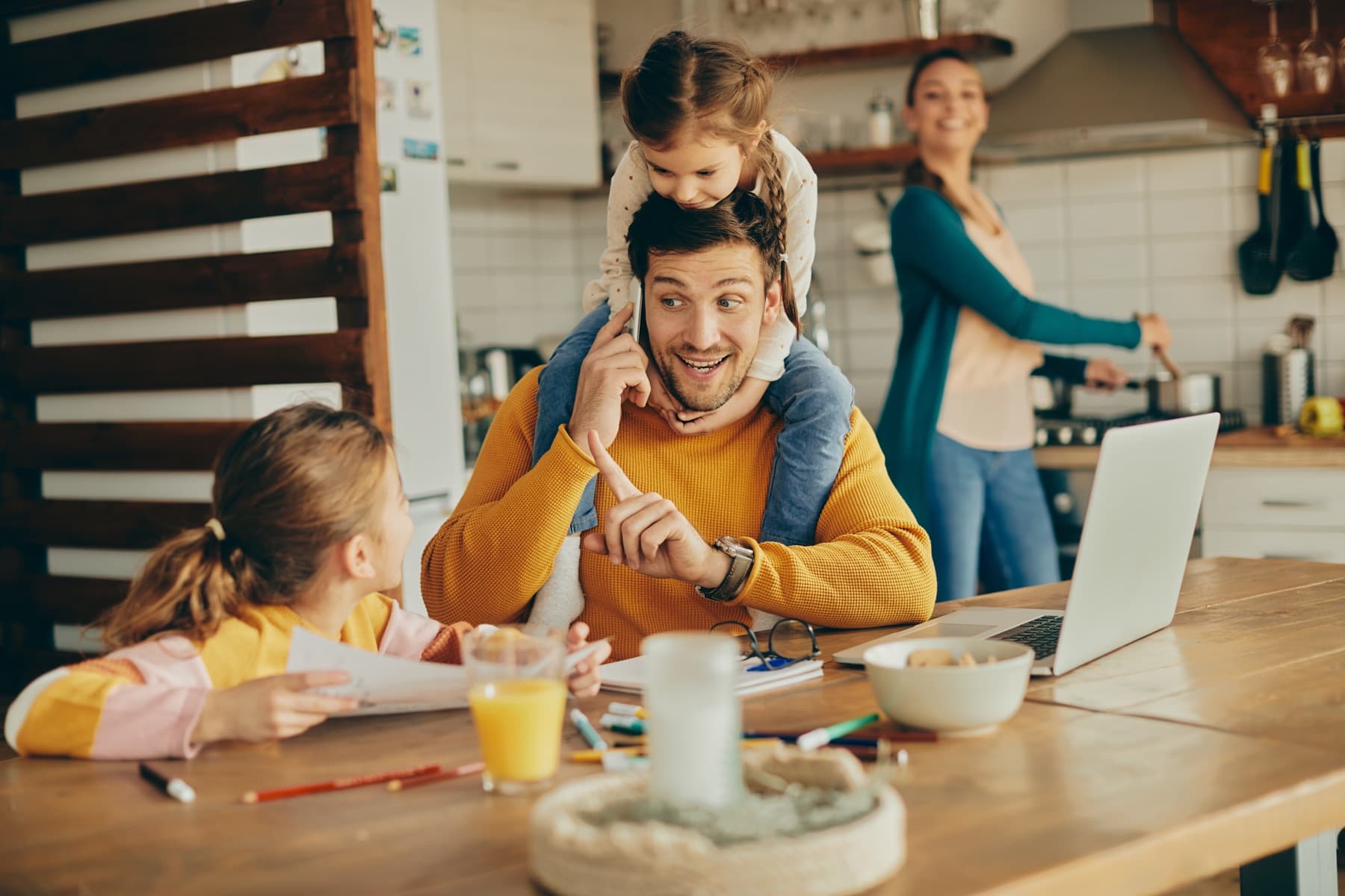 A father on the phone with a child on his shoulders, another child and mother in the kitchen behind him